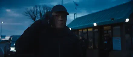 A moody scene from the movie The Amateur (2025) featuring Laurence Fishburne. He is wearing a black cap and jacket, engaged in a phone conversation against an evening sky.