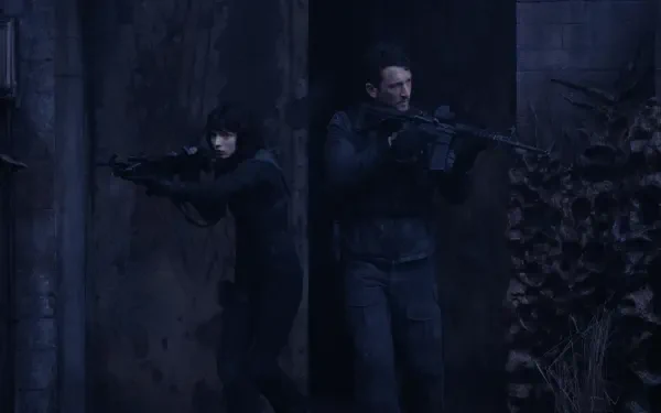 Two characters stealthily move through a shadowy environment, armed with rifles. This tense scene is from the movie *The Gorge* (2025) featuring Miles Teller and Sigourney Weaver.