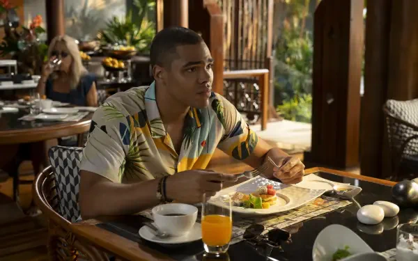A scene from the TV show The White Lotus, featuring Nicholas Duvernay seated at a table, enjoying a meal, with a vibrant background that captures the show's tropical ambiance.