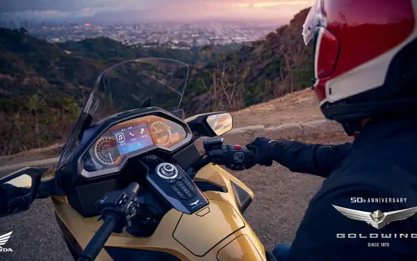 A Honda GL 1800 Gold Wing motorcycle parked on a scenic overlook, with a rider in a red helmet gazing at a vibrant sunset over a cityscape in the background.