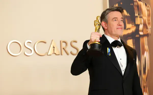 Peter Straughan proudly holds an Oscar trophy, dressed in formal attire, with the iconic OSCARS backdrop. This 4K Ultra HD image captures a moment of celebration at the Academy Awards.