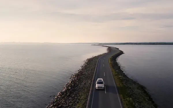A Volvo XC90 Facelift drives along a scenic coastal road, bordered by water, captured in a stunning 4K Ultra HD wallpaper.