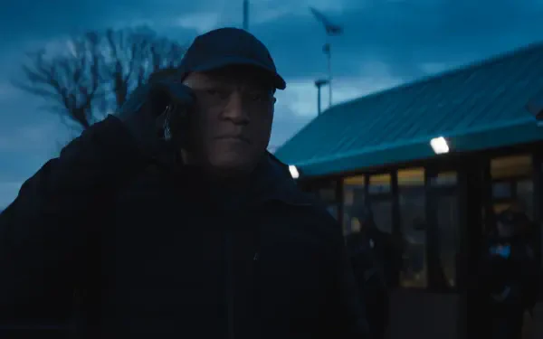 A moody scene from the movie The Amateur (2025) featuring Laurence Fishburne. He is wearing a black cap and jacket, engaged in a phone conversation against an evening sky.