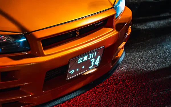 Close-up of an orange Nissan Skyline GT-R R34 front end with illuminated headlights and Japanese license plate, captured in 4K Ultra HD for a PC desktop wallpaper.