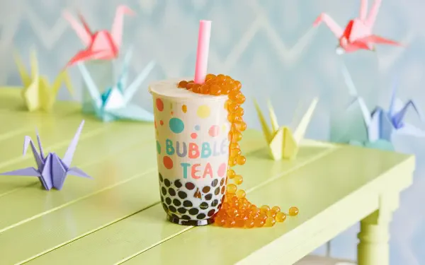 4K Ultra HD desktop wallpaper featuring a colorful bubble tea cup with tapioca pearls and orange popping boba, surrounded by origami cranes on a pastel yellow table.