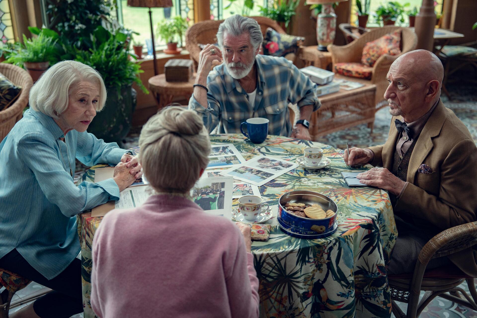 4K Ultra HD PC desktop wallpaper: scene from the movie The Thursday Murder Club — four older friends gathered around a floral table strewn with photos and clues in a cozy lounge.