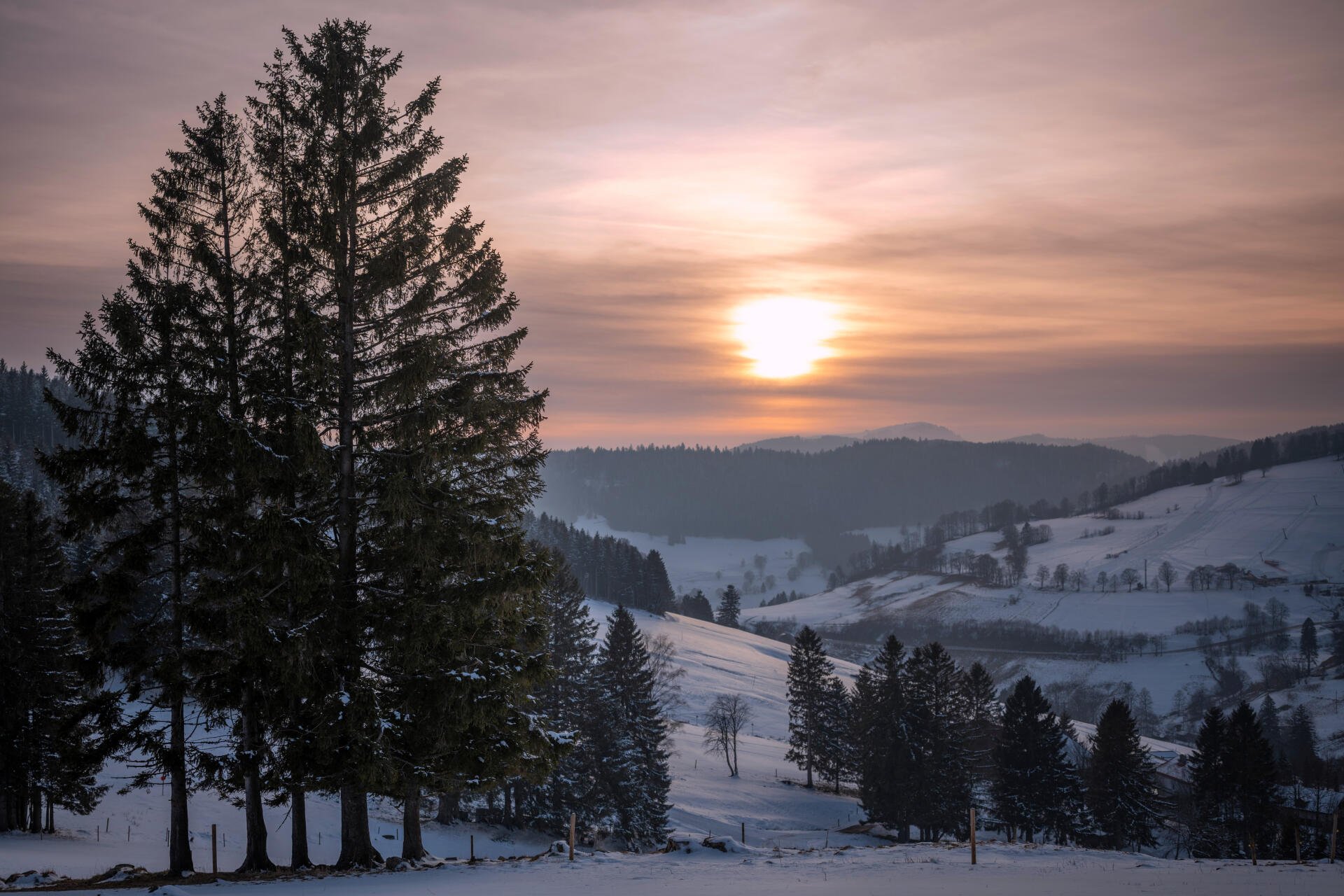 Sunset over a snow-covered Schwarzwald valley in Germany, silhouetted firs in the foreground, rolling hills and distant villages — 4K Ultra HD winter PC desktop wallpaper