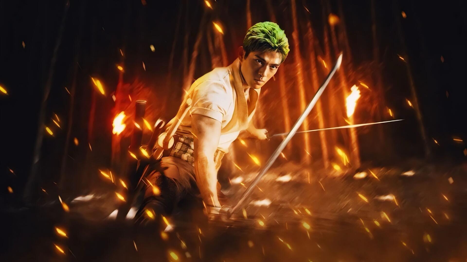 One Piece (Live Action) 2K Quad HD PC desktop wallpaper: green‑haired swordsman crouches with drawn blades amid flying sparks and roaring flames.