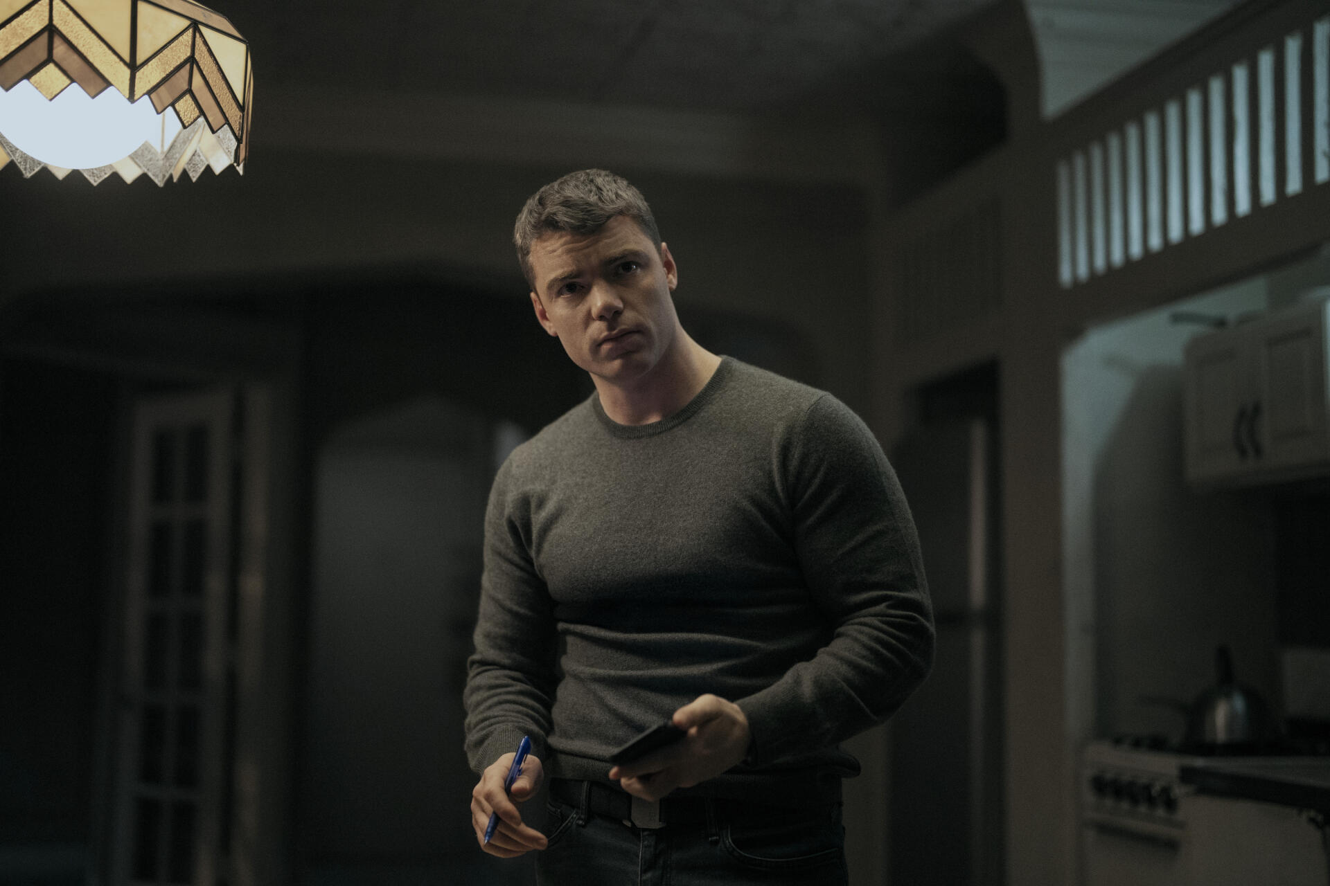 Thriller 2K Quad HD PC wallpaper: brooding man in a dim, shadowed kitchen holding a notebook, tense moody lighting evoking The Night Agent.