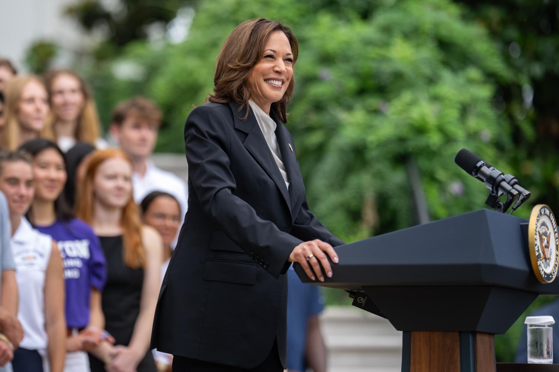 Smiling woman in a dark suit speaking at a podium with a seal, addressing a diverse crowd outdoors — HD desktop wallpaper background.