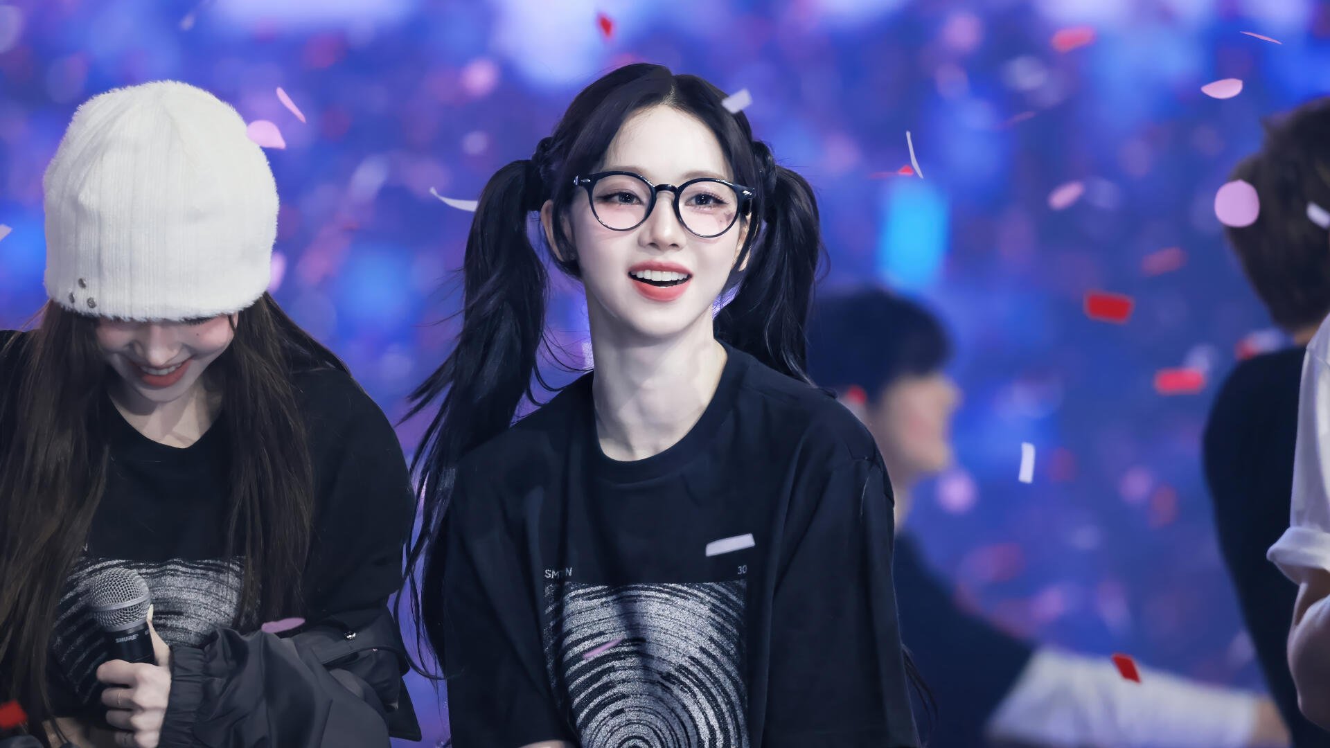 4K Ultra HD desktop wallpaper of a smiling K-pop singer in glasses and pigtails onstage, wearing a black tee as confetti falls against vibrant, blurred concert lights.