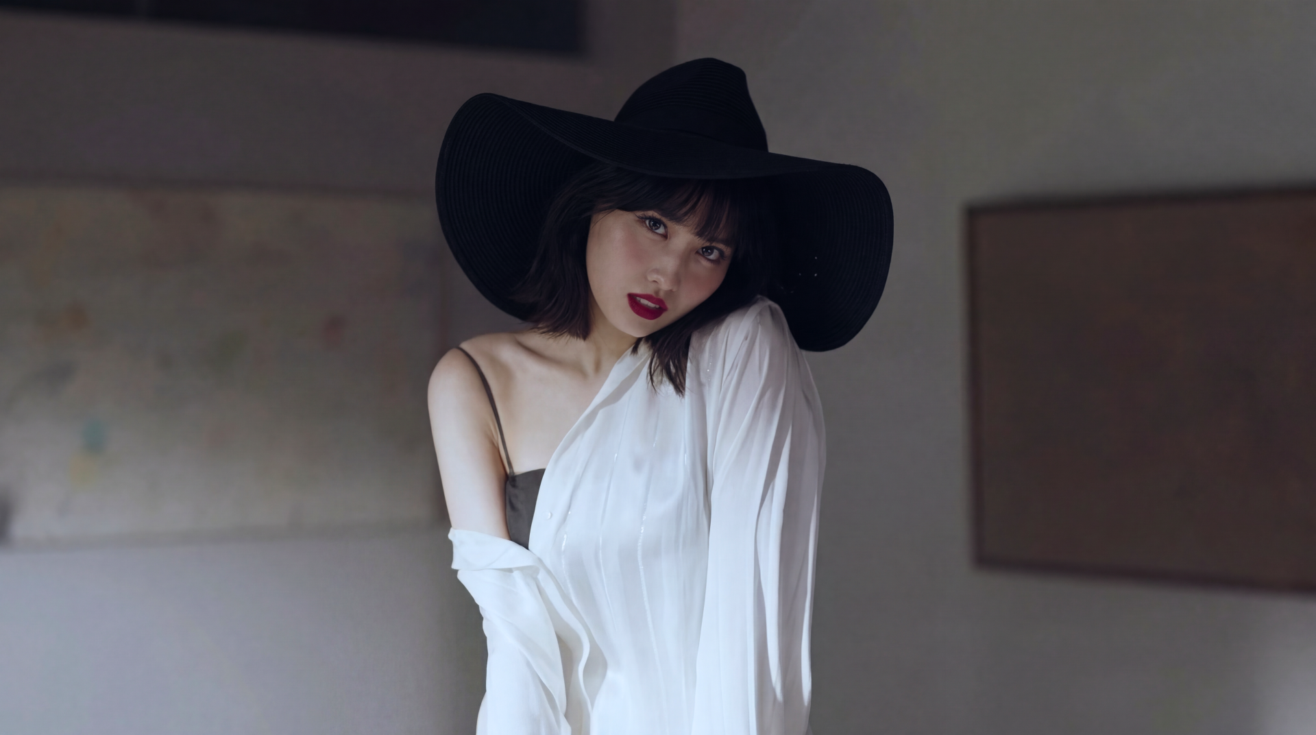 Woman in a wide-brim black hat and white off-shoulder top, gazing toward the camera in a softly lit interior — 5K Ultra HD desktop wallpaper.