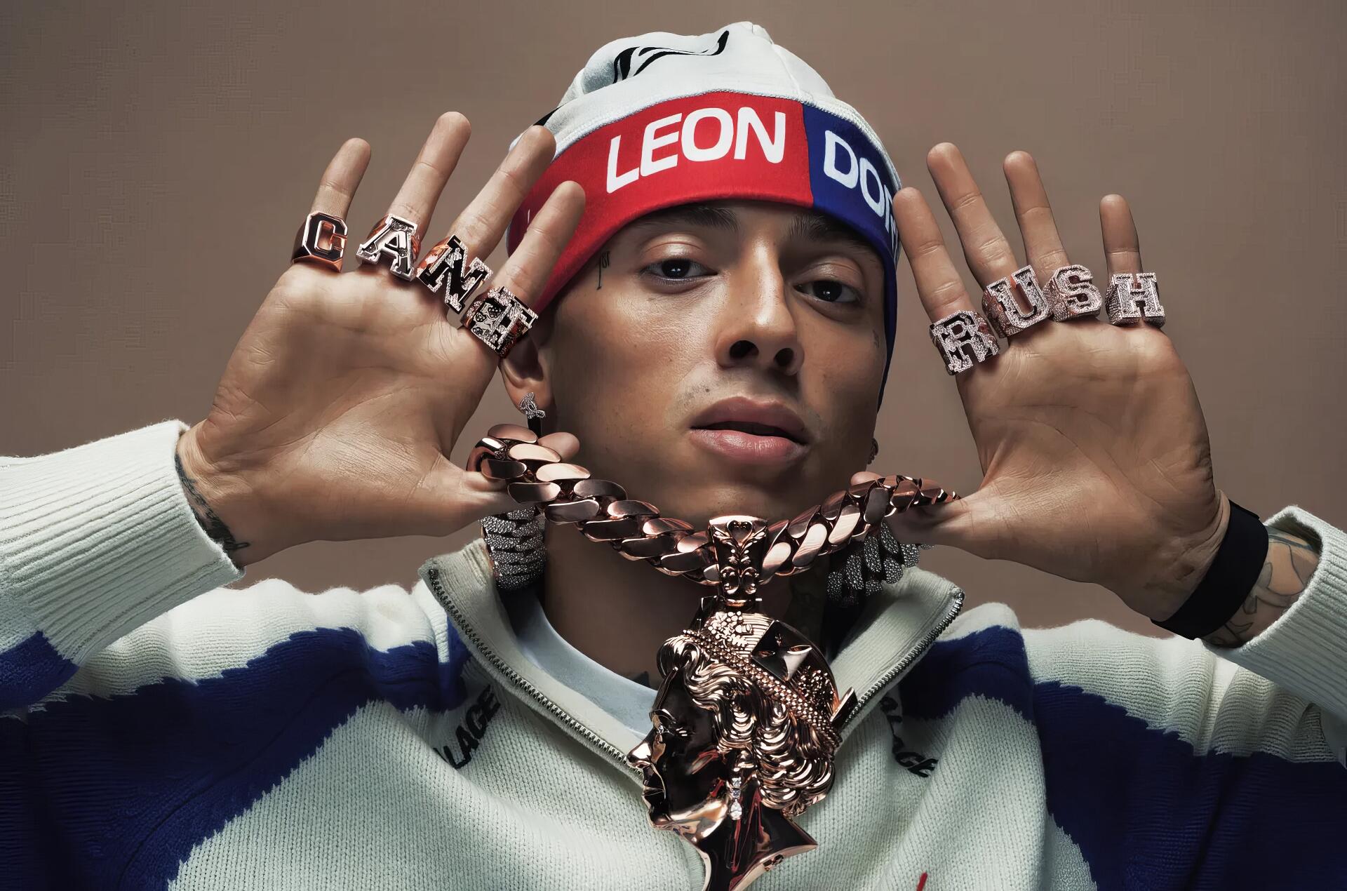 HD PC desktop wallpaper of a person wearing a red-white-blue headband reading LEON, heavy chain pendant and lettered rings, hands raised toward the camera against a beige background.