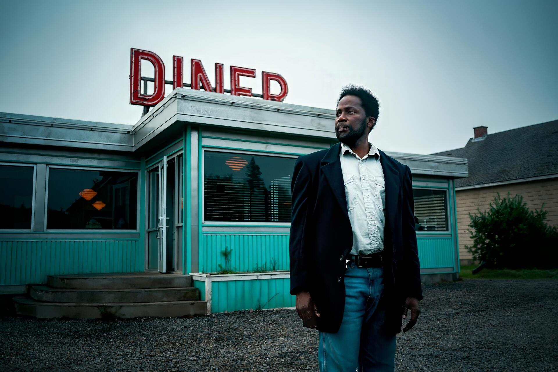 HD PC desktop wallpaper/background from TV show From (2022): a suited man stands before a turquoise roadside diner beneath a moody, overcast sky.