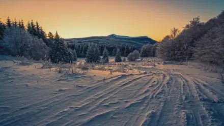 4K Ultra HD PC desktop wallpaper: cold winter landscape at sunset, snow tracks leading toward frosted trees and distant mountains beneath a warm golden-orange sky.