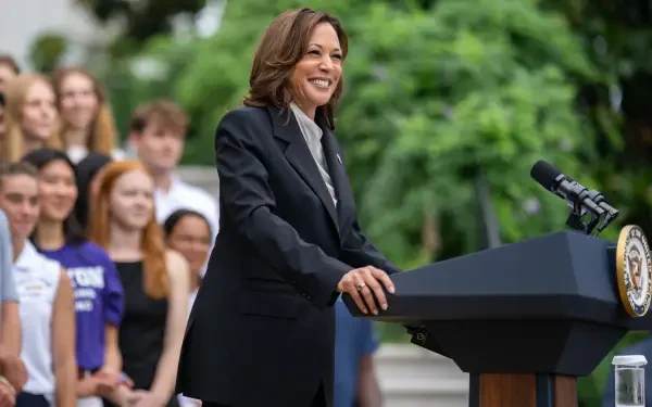 Smiling woman in a dark suit speaking at a podium with a seal, addressing a diverse crowd outdoors — HD desktop wallpaper background.