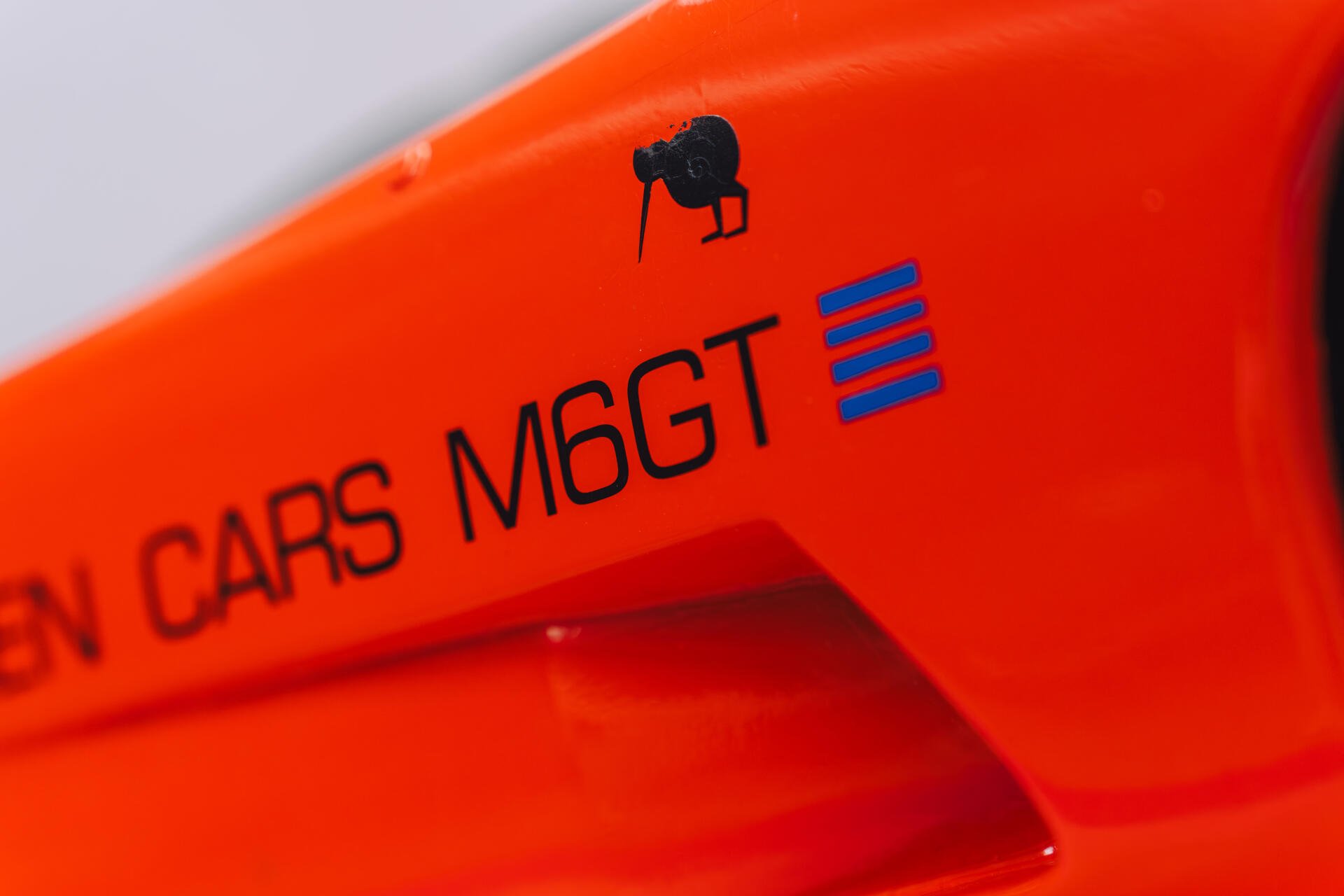 Close-up of bright orange McLaren G6GT vehicle showing the M6GT badge and body detail — 8K Ultra HD PC desktop wallpaper/background.