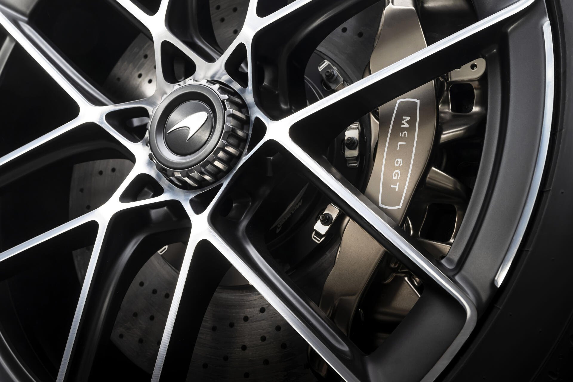 Close-up 8K Ultra HD desktop wallpaper of a McLaren McL 6GT wheel: black multi-spoke rim, McLaren center cap, drilled brake rotor and branded caliper.