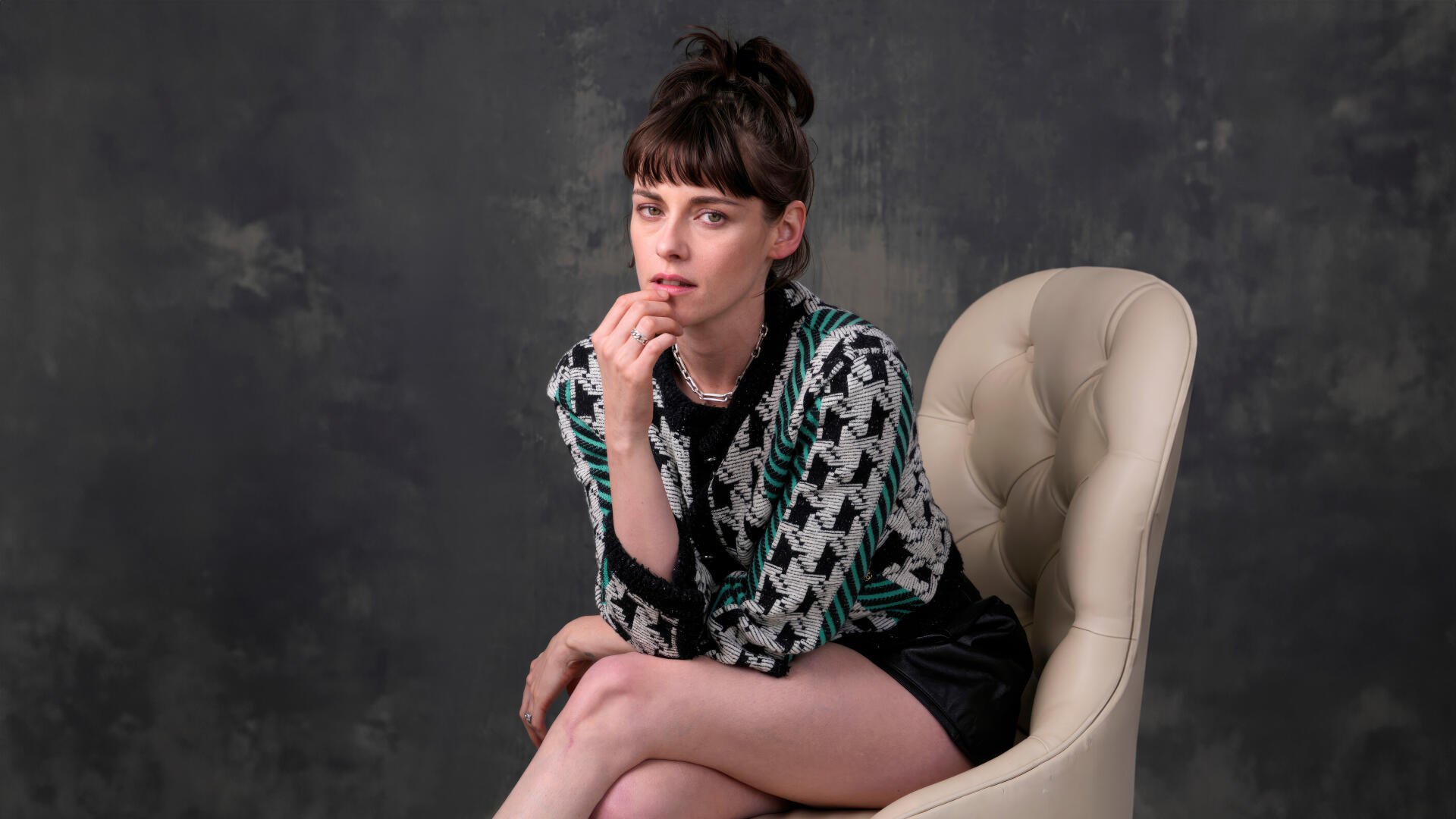 Seated celebrity actress portrait as an 8K Ultra HD PC desktop wallpaper: patterned sweater and shorts, hair up, contemplative pose on a cream chair against a muted studio backdrop.