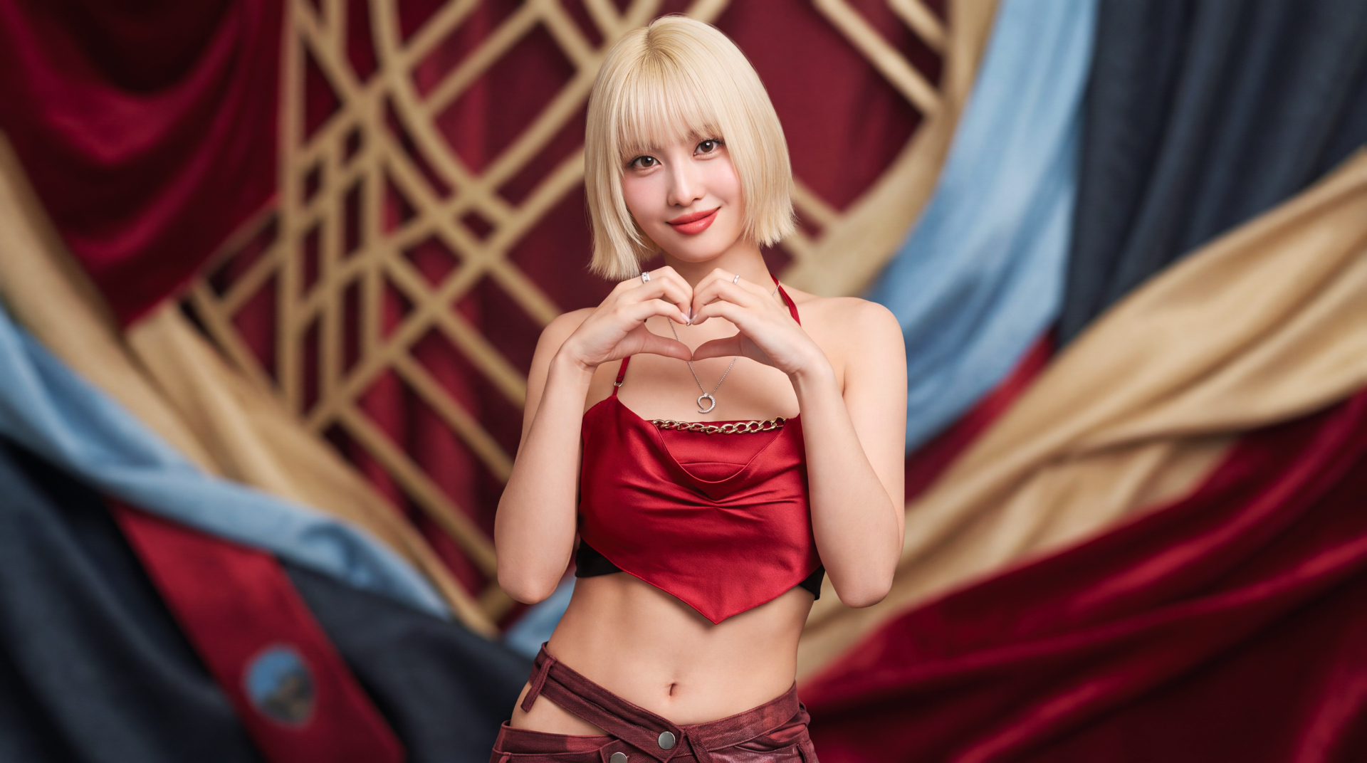 Blonde woman in a red satin crop top forming a heart with her hands against an ornate draped backdrop — 5K Ultra HD PC desktop wallpaper with K-pop–inspired styling.