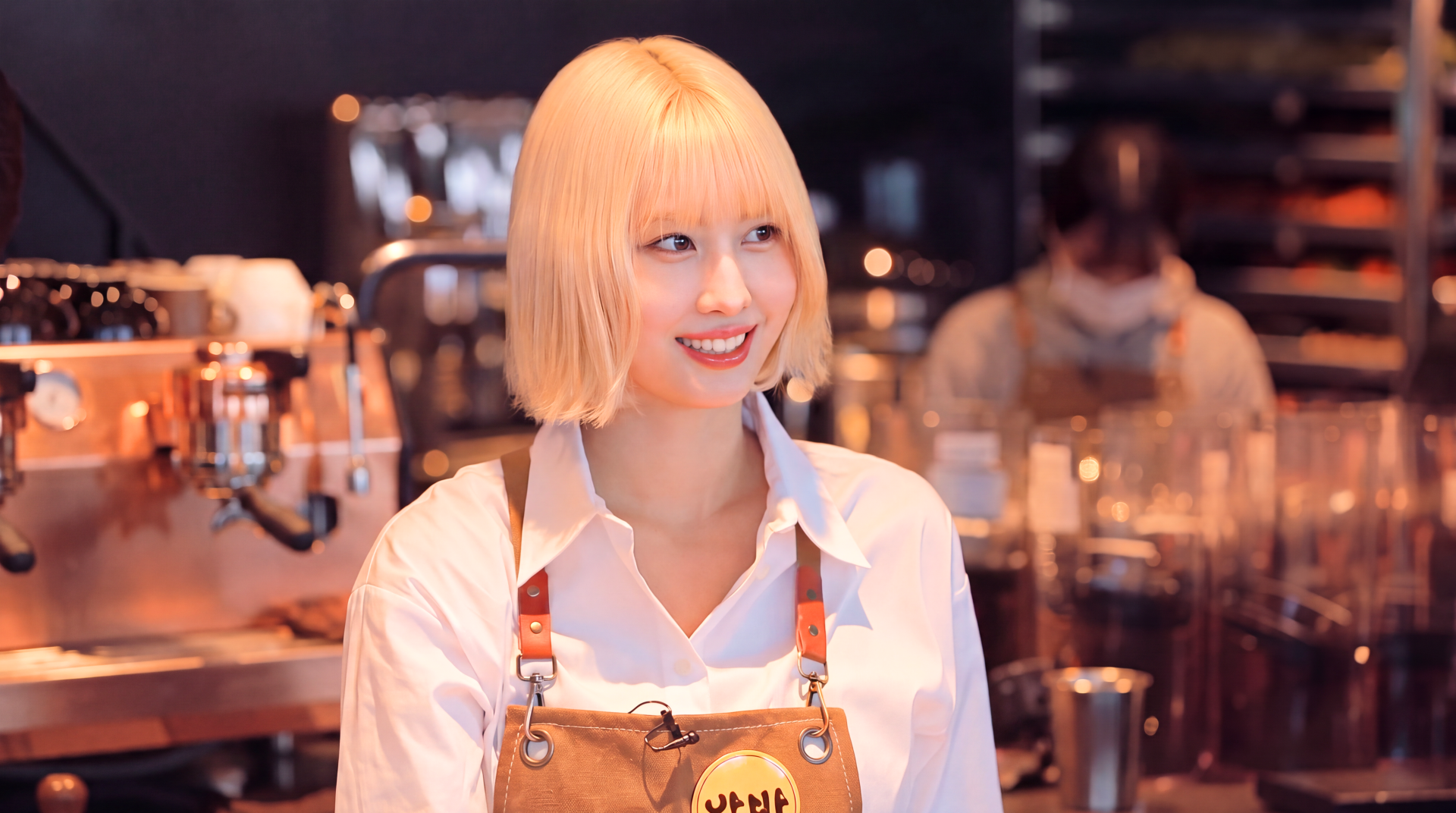 Smiling blonde woman in apron at a café counter with warm bokeh lights — 5K Ultra HD PC desktop wallpaper/background evoking K-pop idol and Japanese model style.