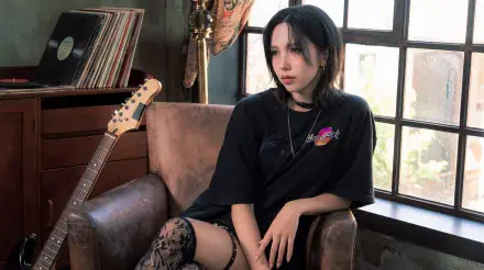 Goth-style woman with dark hair in an oversized black tee and lace stockings sits in an armchair by a window, guitar and vinyl nearby — moody 5K desktop wallpaper.