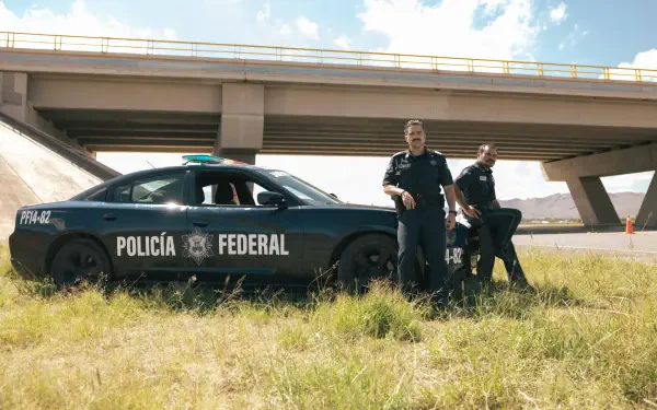 4K Ultra HD PC desktop wallpaper background Facing El Chapo: two Policía Federal officers by a patrol car beneath a highway overpass, sunlit grass and blue sky.