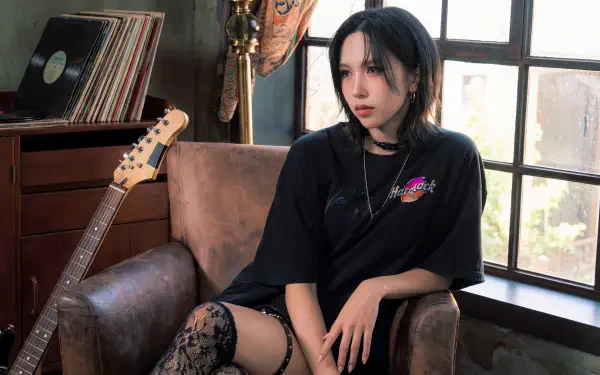 Goth-style woman with dark hair in an oversized black tee and lace stockings sits in an armchair by a window, guitar and vinyl nearby — moody 5K desktop wallpaper.