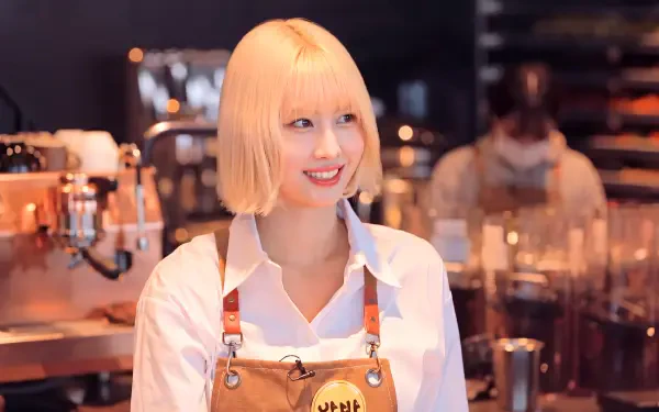 Smiling blonde woman in apron at a café counter with warm bokeh lights — 5K Ultra HD PC desktop wallpaper/background evoking K-pop idol and Japanese model style.