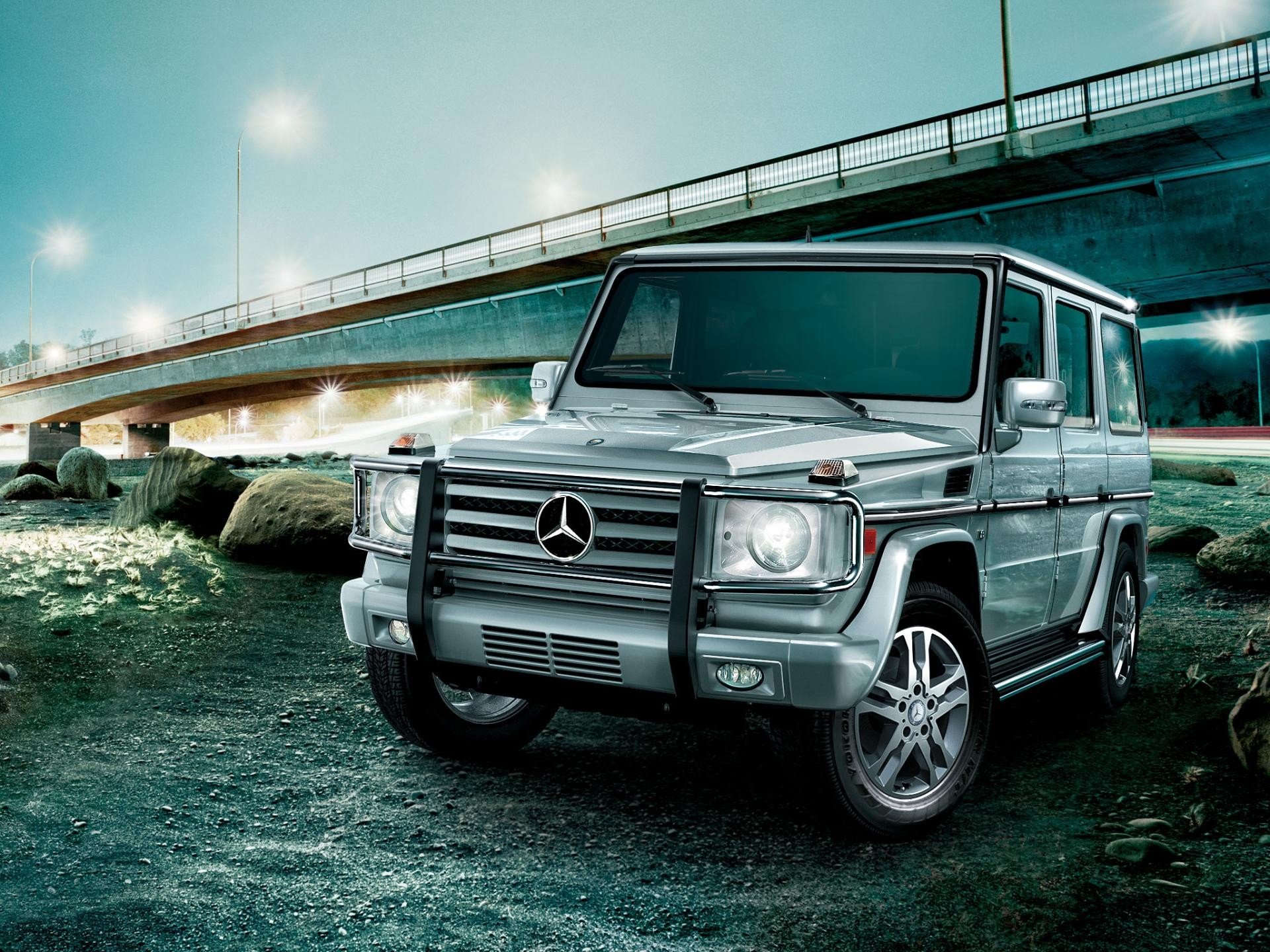 HD PC desktop wallpaper: silver Mercedes G-Class SUV parked by a waterfront beneath a lit highway bridge, moody teal-lit vehicle background.