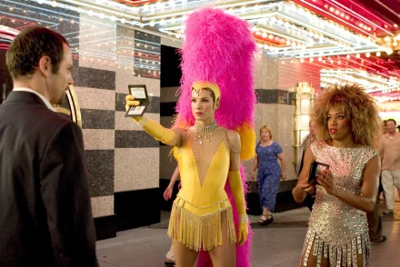 HD PC wallpaper: scene from Miss Congeniality 2: Armed and Fabulous — showgirl in yellow feathered costume flashes a phone under a neon theater marquee while a sequinned companion watches.