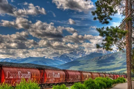 A scenic view of a train in Canada, with vibrant clouds and impressive mountains in the background, creating a stunning HD desktop wallpaper.