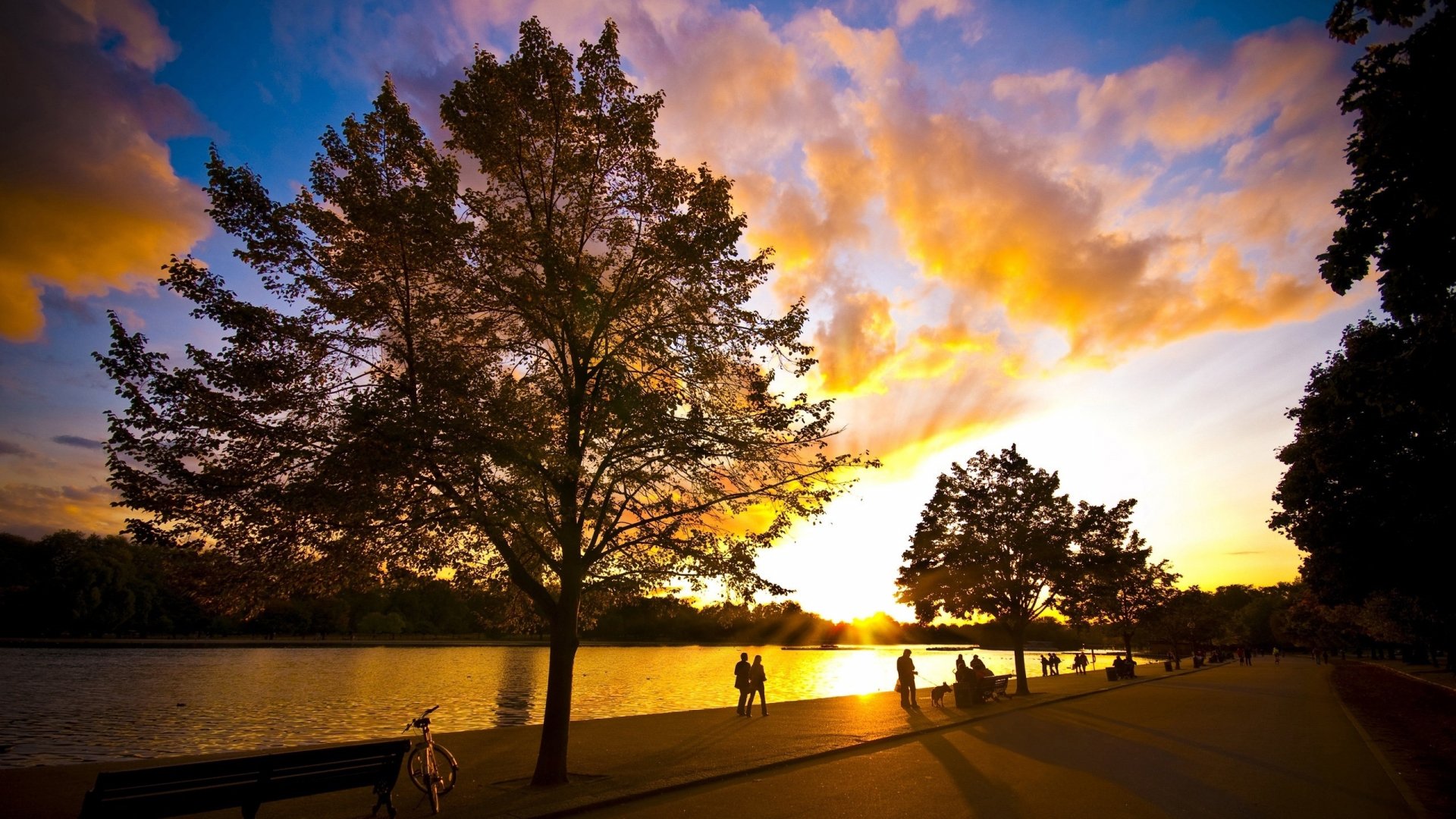 Sunset over a park lake with silhouetted trees and people along the shore, captured in vibrant colors as an HD PC desktop wallpaper background.