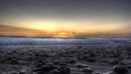 HD PC desktop wallpaper and background: low-angle pebble beach at sunset, wet stones in foreground, rolling waves and a golden-orange horizon beneath a fading blue sky.
