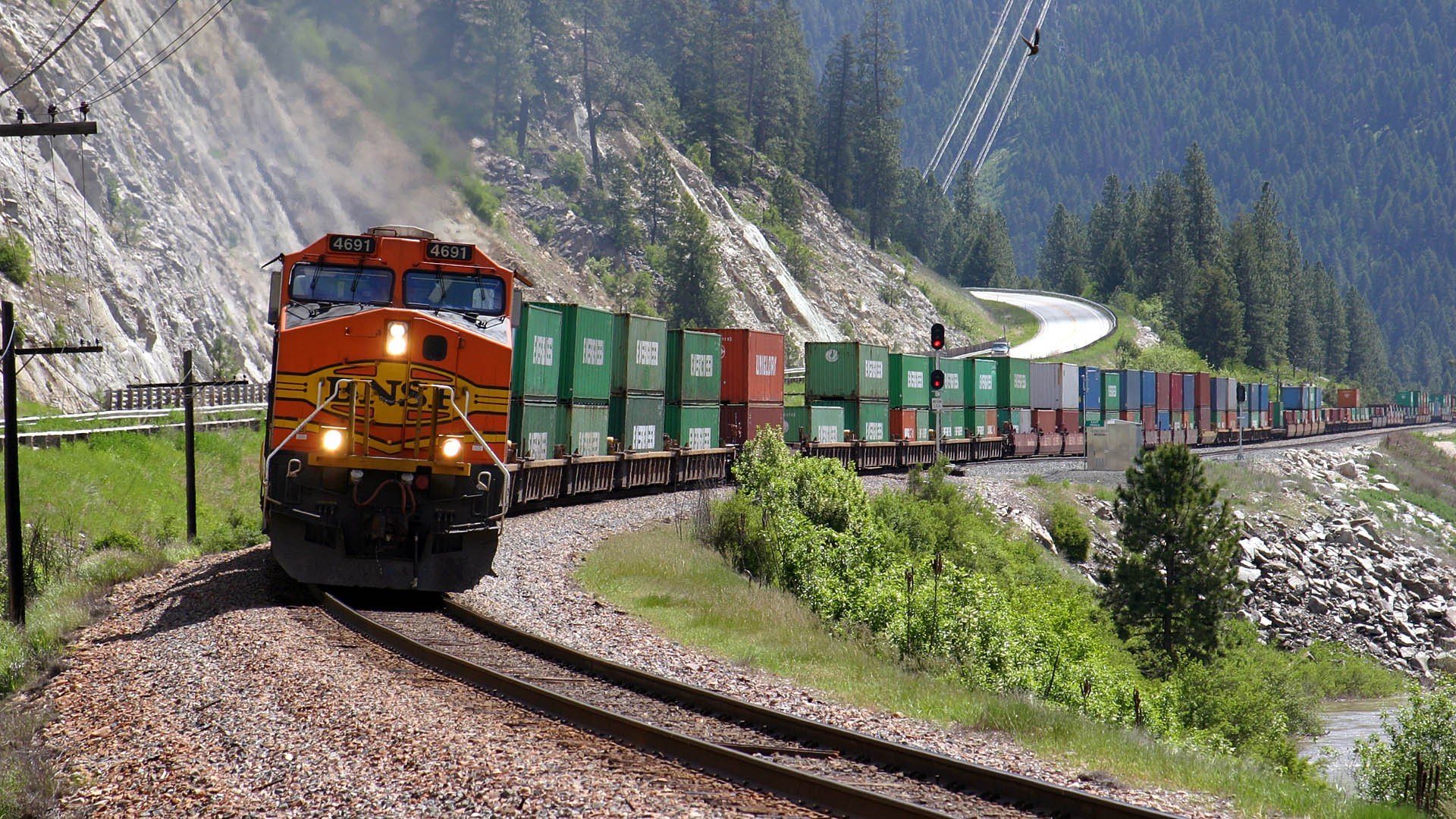 HD desktop wallpaper featuring a freight train winding through a scenic mountainous landscape with lush greenery and steep rocky cliffs.