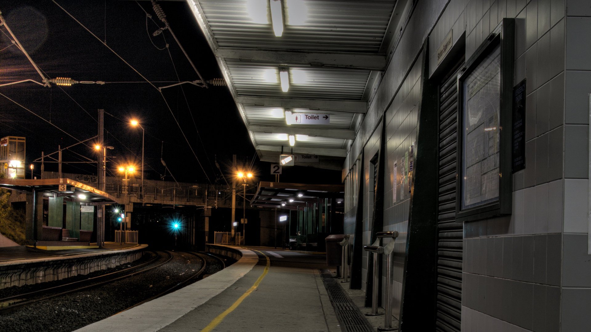 HD PC desktop wallpaper and background: nighttime man-made railroad station platform lit by fluorescent lights, curved tracks vanishing into a dark tunnel, empty sheltered waiting area.