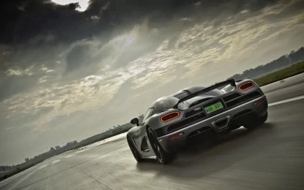 HD desktop wallpaper featuring a Koenigsegg Agera speeding down a wet road under a dramatic, cloud-filled sky. The sleek car showcases its impressive design and power.