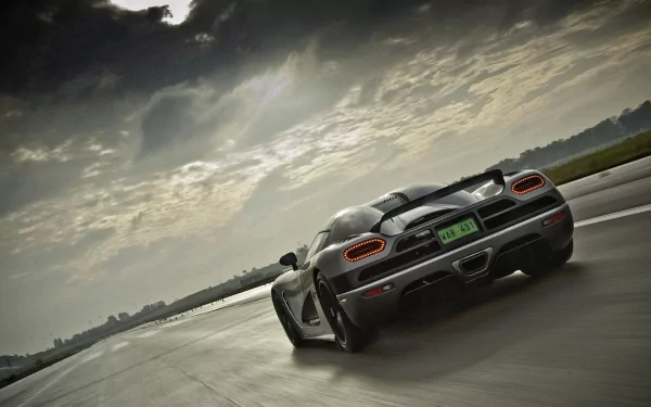 HD desktop wallpaper featuring a Koenigsegg Agera speeding down a wet road under a dramatic, cloud-filled sky. The sleek car showcases its impressive design and power.