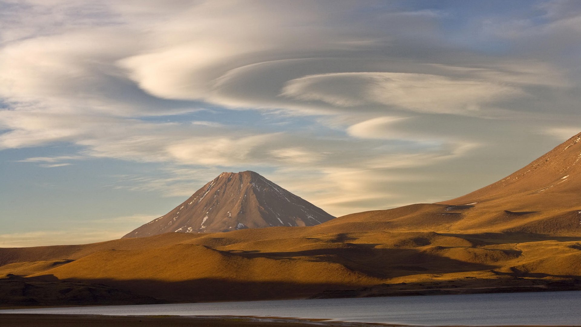 HD PC desktop wallpaper featuring a volcano rising above rolling hills under a sky with swirling cloud formations in a serene natural landscape.