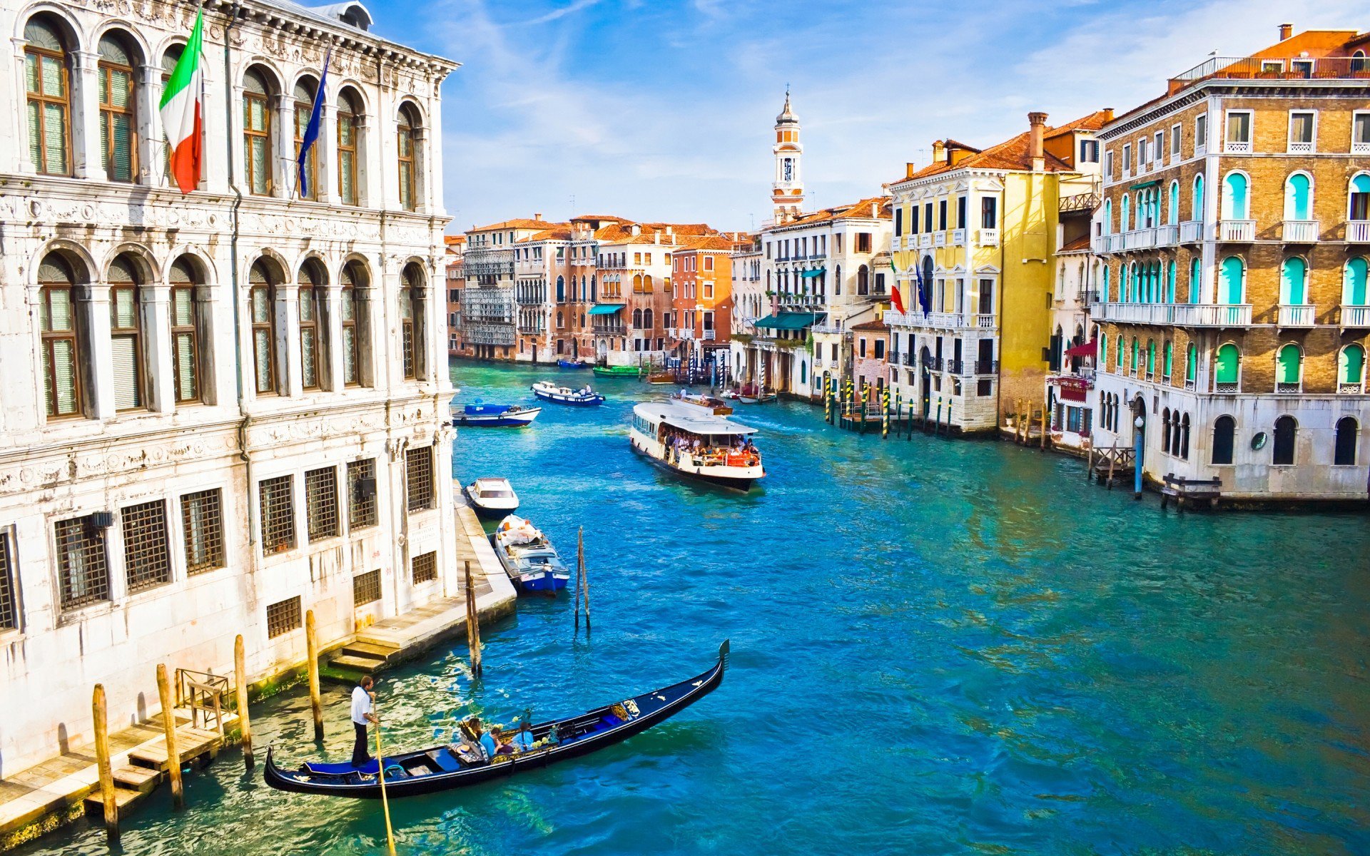 A vibrant HD desktop wallpaper of Venice, Italy, featuring colorful buildings, a bustling canal with boats, and classic man-made architecture along the waterway.
