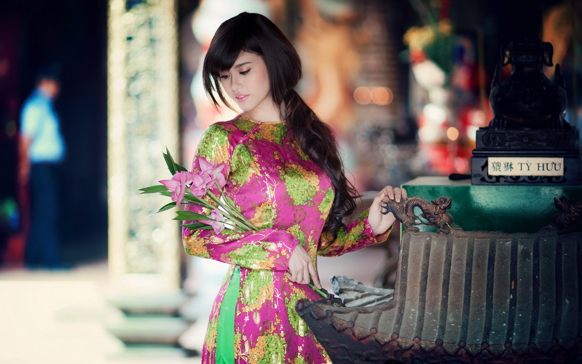 HD desktop wallpaper featuring an Asian woman in a vibrant pink and green dress, holding a bouquet of flowers in a beautifully adorned, traditional setting.