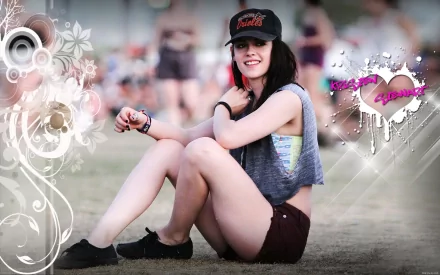 HD desktop wallpaper featuring Kristen Stewart sitting casually on the grass. The background includes artistic floral designs and a heart with graffiti-style text.