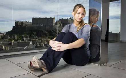 Celebrity Jennifer Lawrence sitting on the floor next to a reflective wall, with a scenic background of an old castle. This HD desktop wallpaper captures a serene and thoughtful moment.