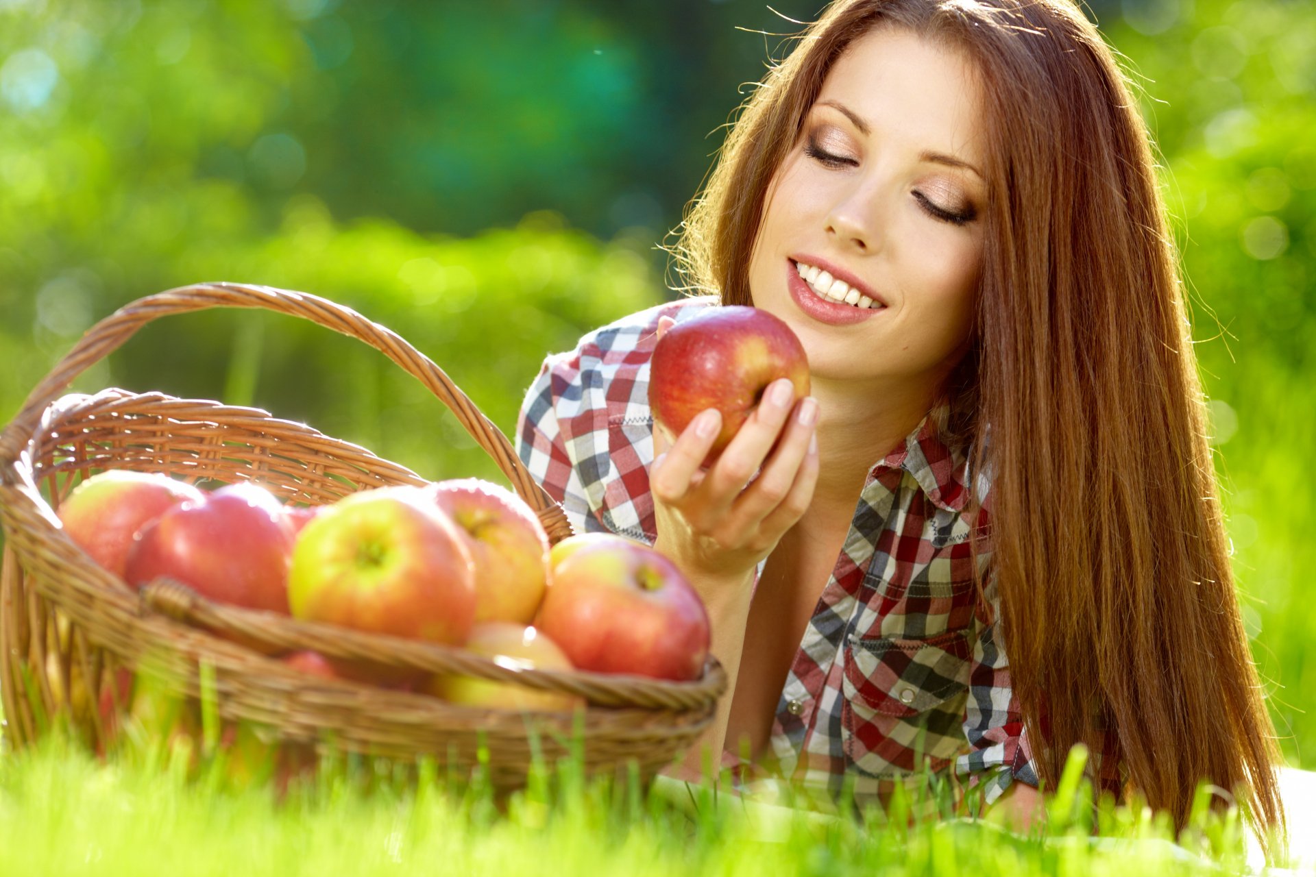 A woman with long hair smiling gently while holding an apple near a basket of apples, set in a vibrant outdoor scene. 4K Ultra HD PC desktop wallpaper.