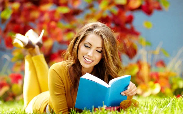 A stylish woman enjoys a book while lounging on the grass, surrounded by vibrant fall colors, creating a warm and inviting mood in this 4K Ultra HD wallpaper.