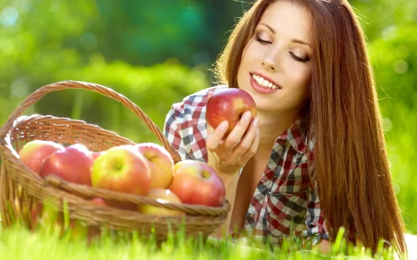 A woman with long hair smiling gently while holding an apple near a basket of apples, set in a vibrant outdoor scene. 4K Ultra HD PC desktop wallpaper.