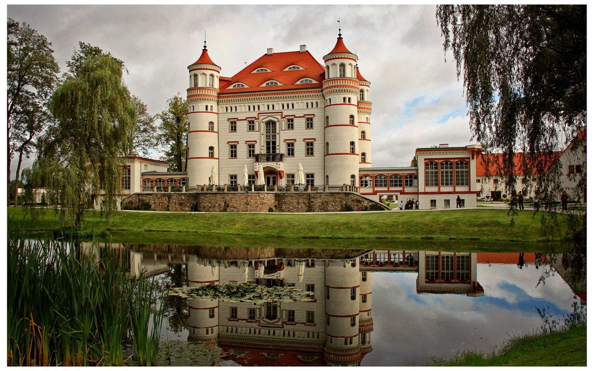 A serene pond reflects a striking man-made castle in Schildau, Poland, surrounded by lush greenery and a tranquil atmosphere, showcasing picturesque architecture.