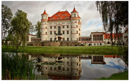 A serene pond reflects a striking man-made castle in Schildau, Poland, surrounded by lush greenery and a tranquil atmosphere, showcasing picturesque architecture.