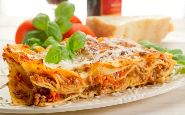 A delicious serving of pasta lasagna garnished with fresh basil, accompanied by tomatoes and bread, showcased as an HD PC desktop wallpaper and background.