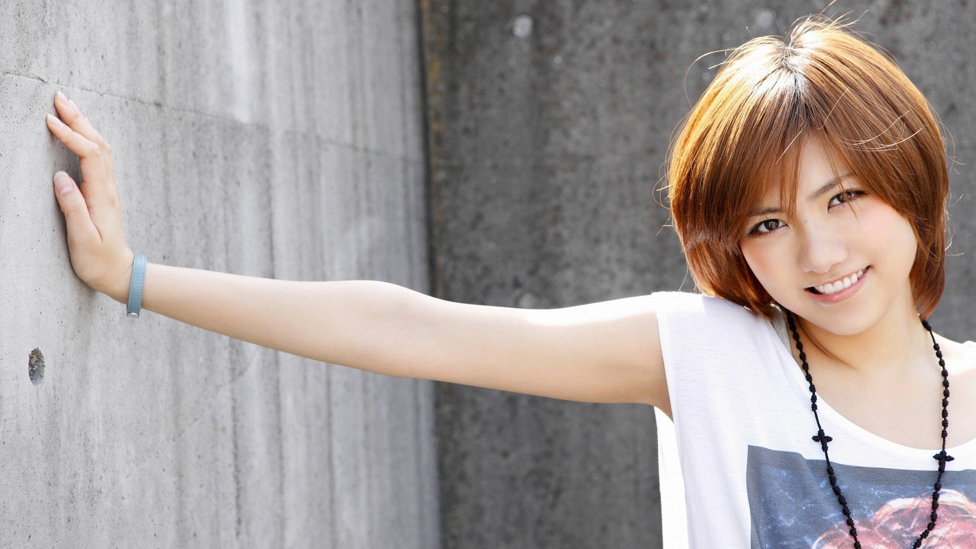 HD wallpaper featuring an Asian woman with short hair, smiling and leaning against a concrete wall background.